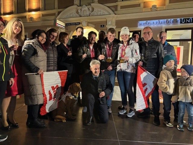 A group of people, including men, women, and children, pose for a photo with medals and wine glasses. They are standing and kneeling in front of a building with flags.