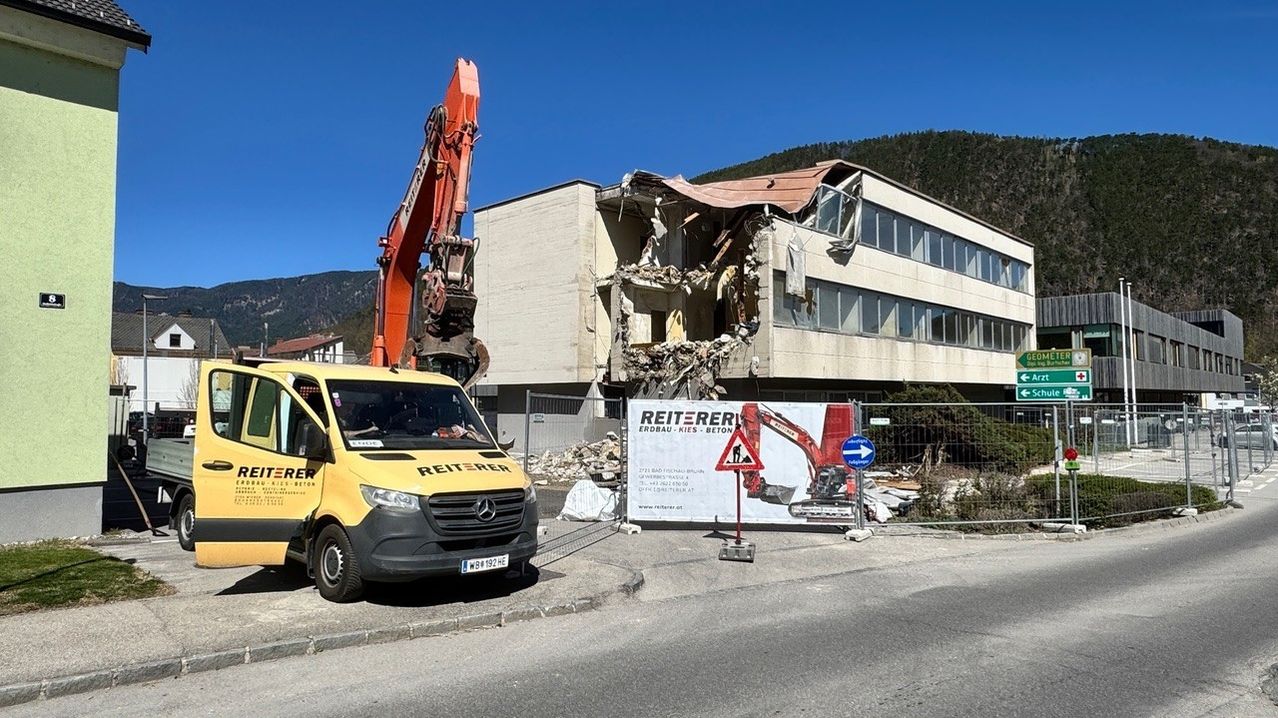 A yellow van with the word Reiterer parked on the side of the road. An excavator is demolishing a building with a sign in front. Mountains are in the background.