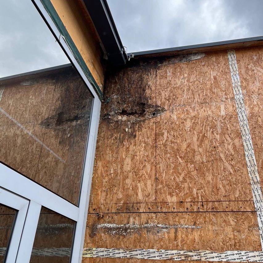 A section of a building's exterior wall shows extensive damage with large holes and brown stains. Adjacent to it, a window reflects the cloudy sky.
