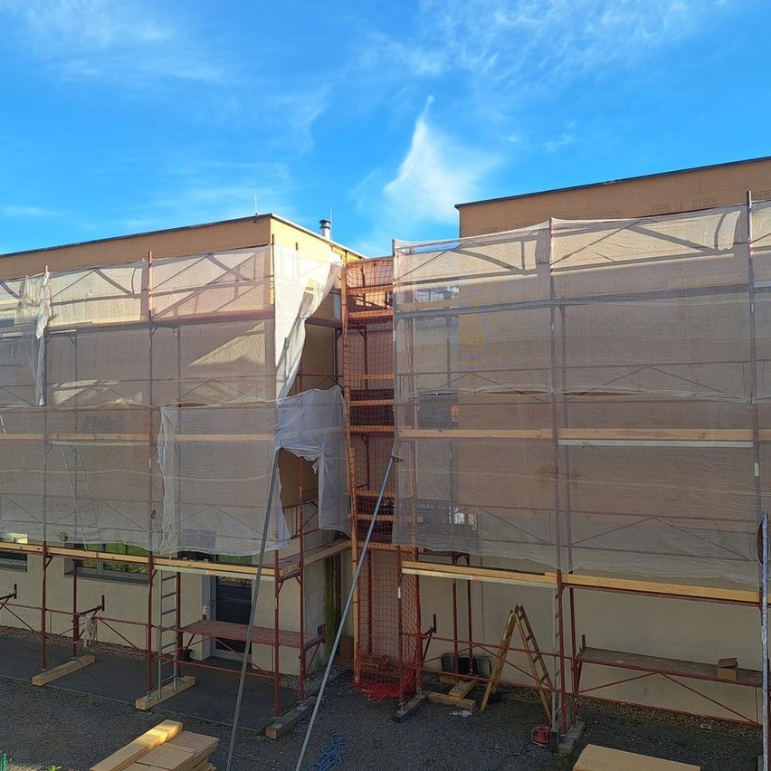A building under construction with scaffolding and netting in place. A clear blue sky is visible in the background.