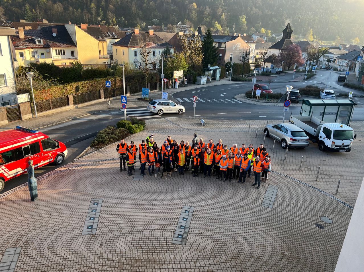 A group of people in orange vests stands in a roundabout. Behind them are houses and trees, with parked cars on the sides. The sky is clear and bright.