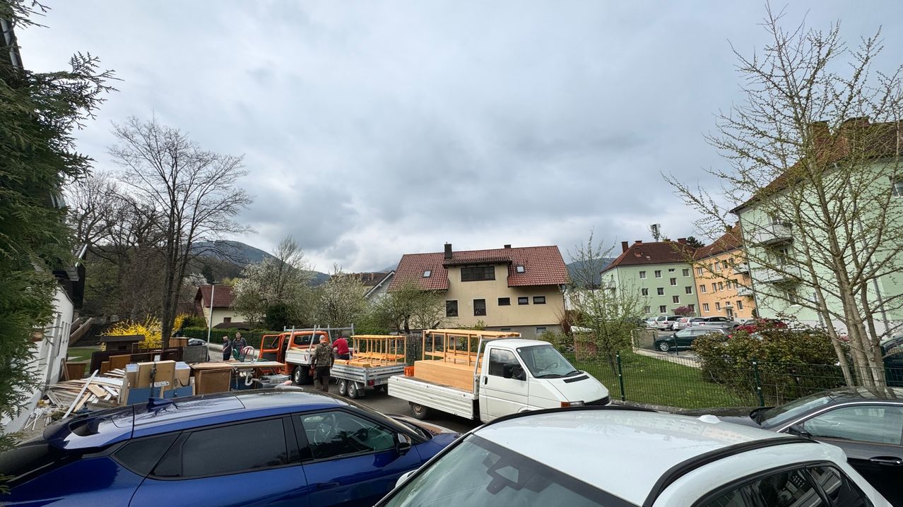 Ein wolkiger Tag in einer Stadt. Zwei Lastwagen sind geparkt, einer hat eine Holzstruktur darauf. Menschen stehen in der Nähe der Lastwagen. Ein Haus mit rotem Dach ist sichtbar.