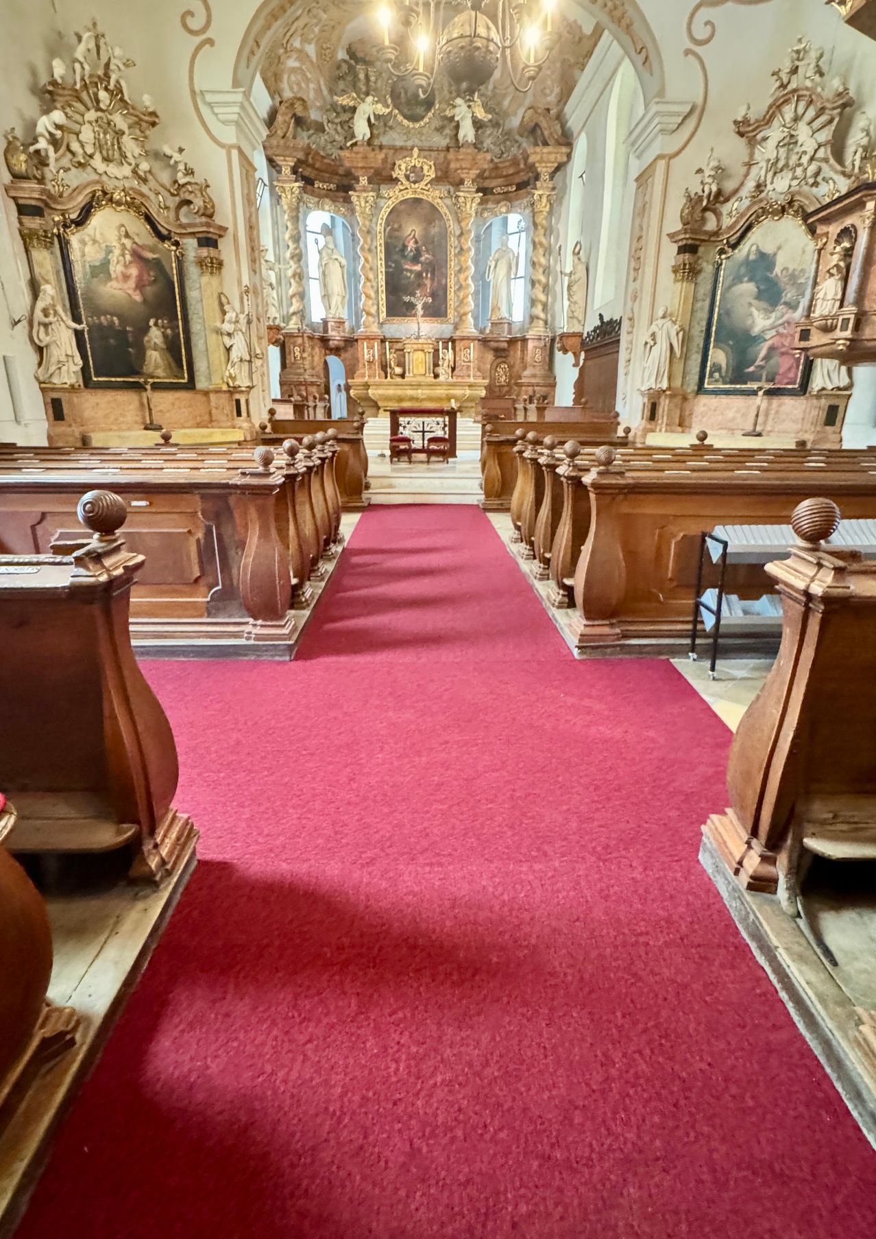 Eine leere Kirche mit hölzernen Kirchenbänken, einem roten Teppich auf dem Gang und zwei großen Gemälden an der Wand.