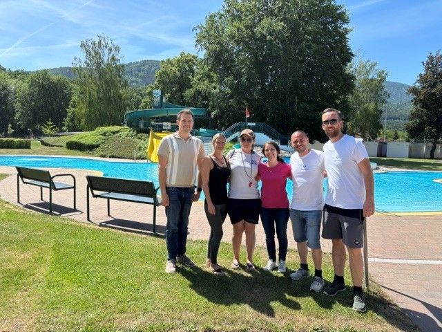 Fünf Personen posieren für ein Foto vor einem Schwimmbad mit einer Wasserrutsche an einem sonnigen Tag.