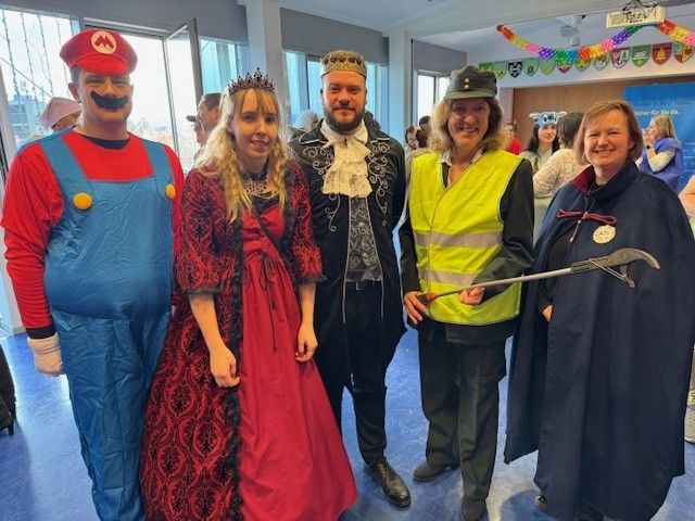 Eine Gruppe von Menschen in Kostümen, darunter ein Mario-Kostüm, eine Krone und ein Besen, posiert für ein Foto in einem Saal.