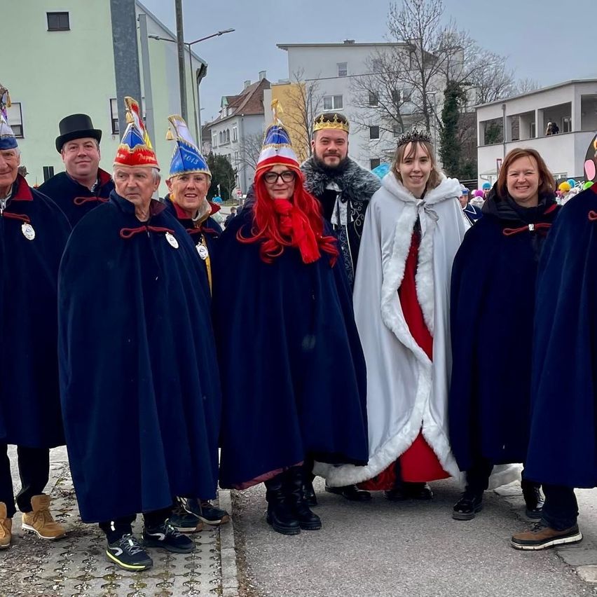 Eine Gruppe von Menschen in festlichen Kostümen steht auf einer Straße. Sie tragen Kronen, Umhänge und Stiefel. Einige tragen Brillen und halten rote Bänder. Bäume und Gebäude sind im Hintergrund.