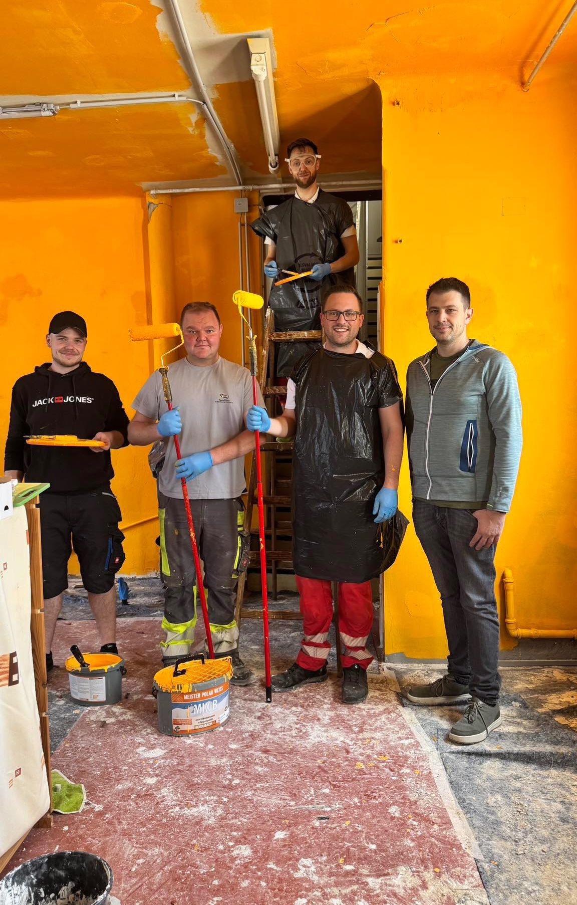 Fünf Männer stehen in einem Raum mit gelben Wänden, halten Farbwalzen und Eimer in der Hand. Sie tragen alle Handschuhe und scheinen sich darauf vorzubereiten, zu malen.