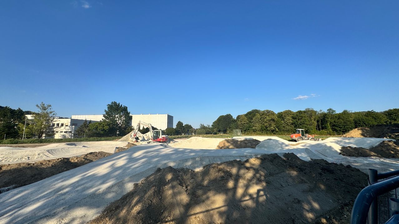A construction site with two excavators, piles of dirt, and a building in the background under a clear blue sky.