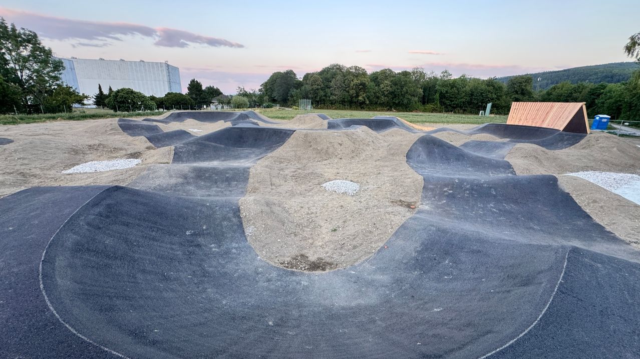 Ein Outdoor-Skatepark mit verschiedenen Rampen und einer zentralen Erdaufschüttung, umgeben von Bäumen und einem Gebäude in der Ferne. Der Himmel ist klar mit einem Hauch von Rosa.