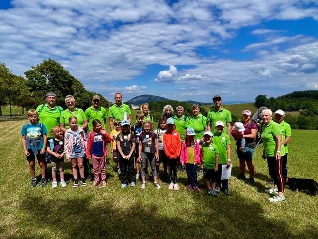 Eine Gruppe von Menschen, einschließlich Erwachsener und Kindern, posiert für ein Foto auf einer Wiese mit einem bewölkten Himmel und Bergen im Hintergrund.