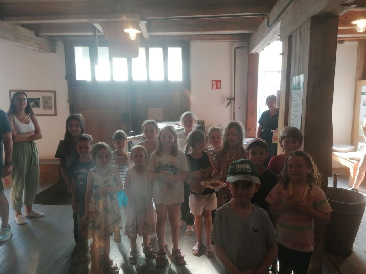 Eine Gruppe von Kindern und Erwachsenen steht in einem Raum mit Holzbalken, einige halten Teller mit Essen. Eine Frau steht im Hintergrund in der Nähe einer Tür.