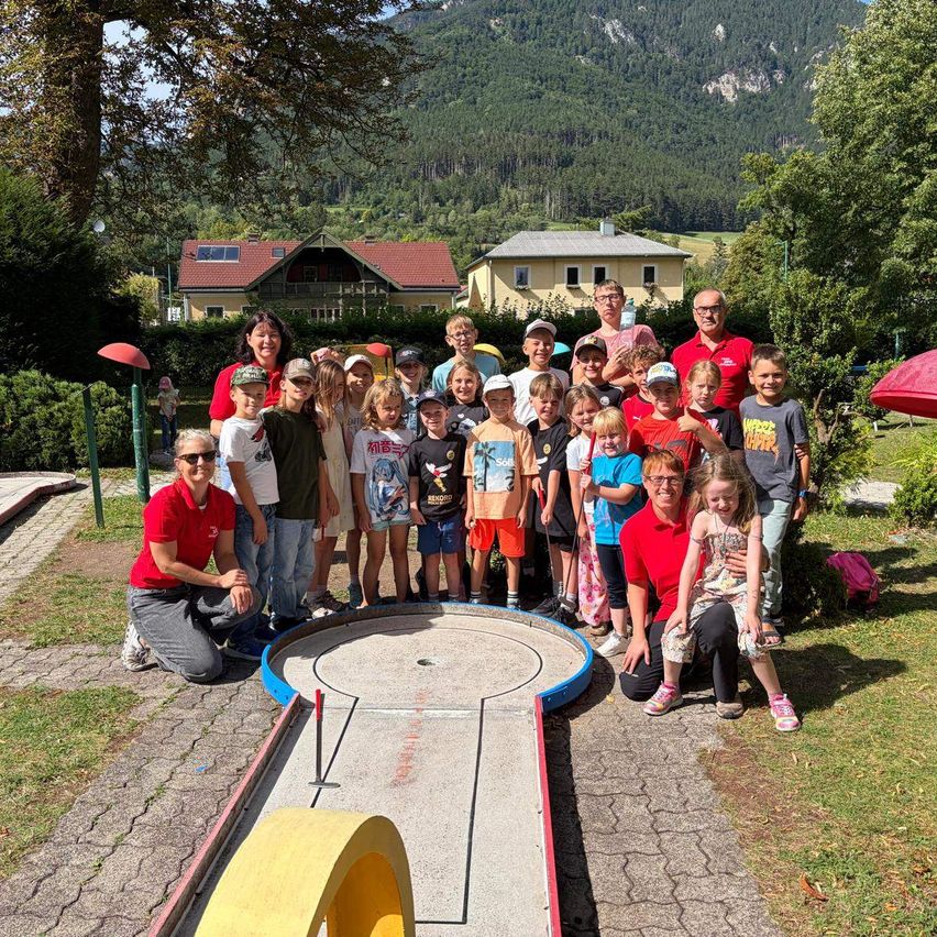 Eine Gruppe von Kindern und Erwachsenen versammelt sich um einen Minigolfplatz, mit Bergen im Hintergrund.