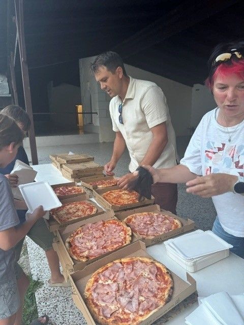 Eine Gruppe von Menschen bereitet sich darauf vor, Pizzas mit Schinken zu essen. Ein Mann in einem weißen Hemd schneidet eine Pizza, während eine Frau mit rosa Haaren zusieht.