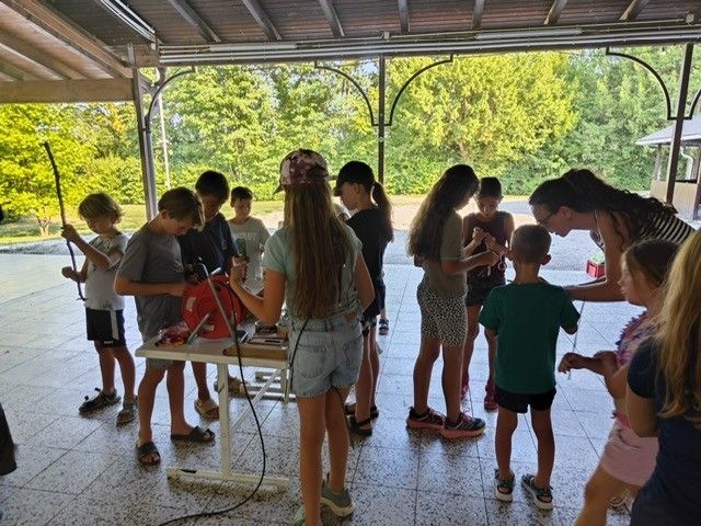 Eine Gruppe von Kindern und Erwachsenen versammelt sich in einem überdachten Bereich, möglicherweise für einen Workshop. Einige halten Werkzeuge und andere beobachten. Eine Frau beugt sich über einen Jungen.