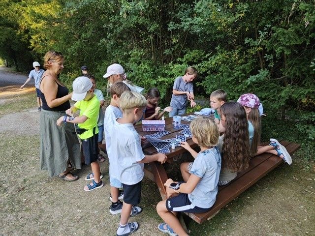 Eine Gruppe von Kindern und Erwachsenen versammelt sich um einen Picknicktisch im Wald und beschäftigt sich mit einer Aktivität. Einige sitzen auf dem Tisch, während andere in der Nähe stehen.