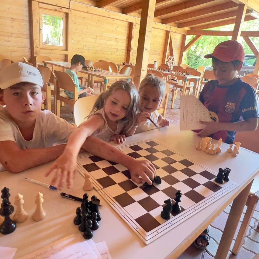 Vier Kinder spielen Schach an einem Tisch. Ein Kind hält ein Schachstück. Der Tisch hat ein Schachbrett und Schachfiguren.