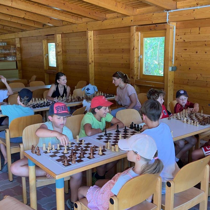 Eine Gruppe von Kindern und Erwachsenen spielt Schach in einem Raum mit Holzwänden und Tischen. Die Kinder sitzen um die Tische herum und sind in ihre Spiele vertieft.