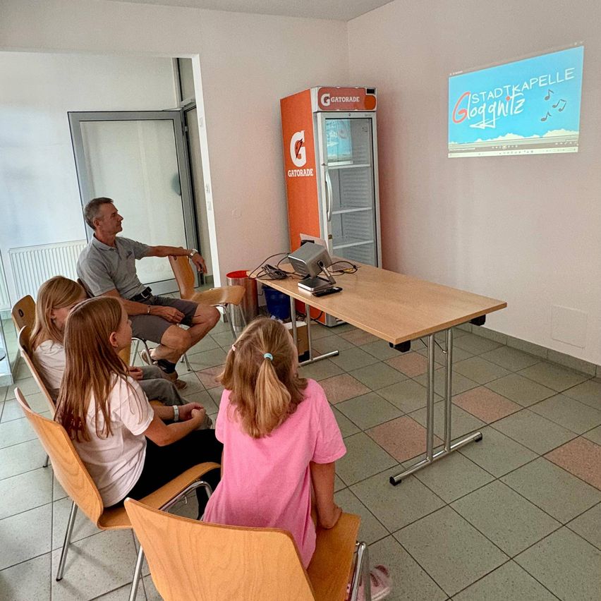 Ein Mann und vier Mädchen sitzen in einem Raum mit einem Tisch, einer Projektionsleinwand und einem Kühlschrank. Der Mann sitzt auf einem Stuhl, und die Mädchen sitzen vor ihm auf Stühlen. Die Projektionsleinwand zeigt eine Präsentation an.