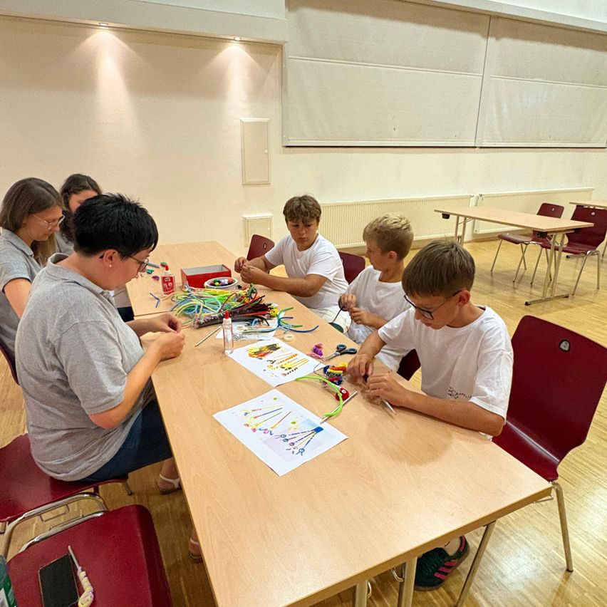 Eine Gruppe junger Menschen sitzt um einen Tisch herum und beschäftigt sich mit einer Bastelaktivität mit verschiedenen bunten Materialien und Werkzeugen. Sie befinden sich in einem Klassenzimmer mit Holzböden und weißen Wänden.