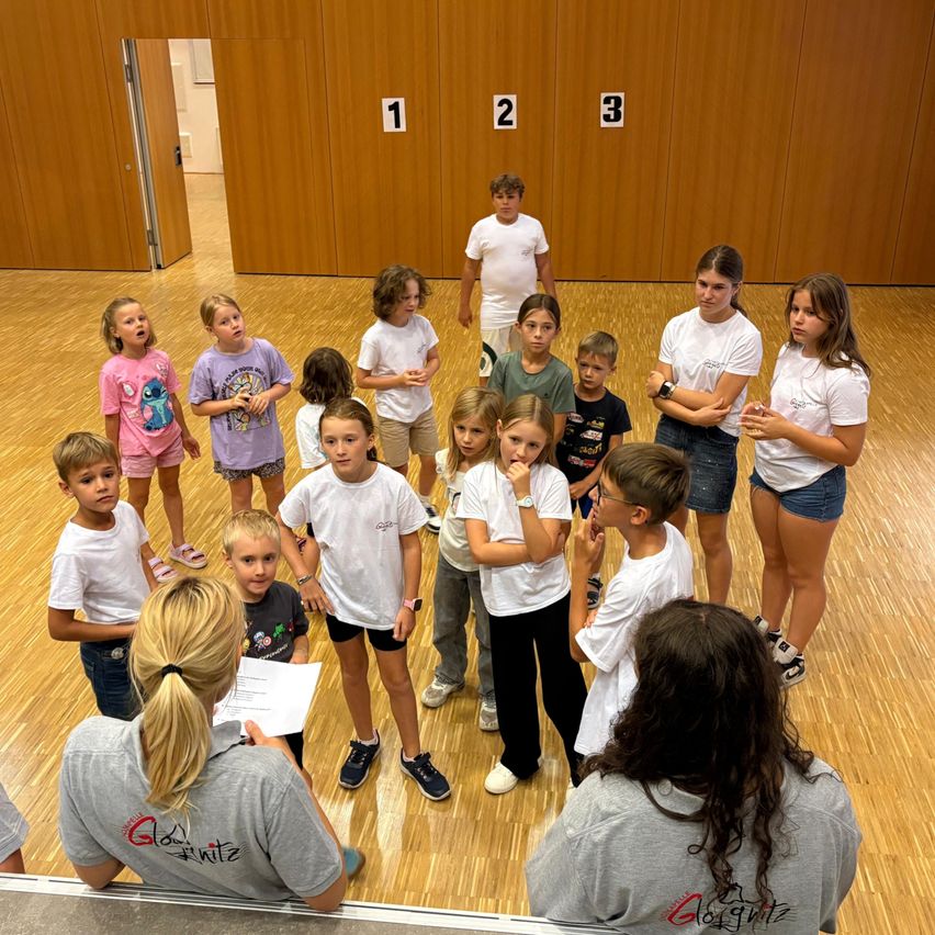 Eine Gruppe von Kindern in einer Turnhalle, einige in weißen T-Shirts, andere in Freizeitkleidung, stehen im Kreis um zwei Erwachsene, einer hält ein Papier und der andere trägt eine Uhr.