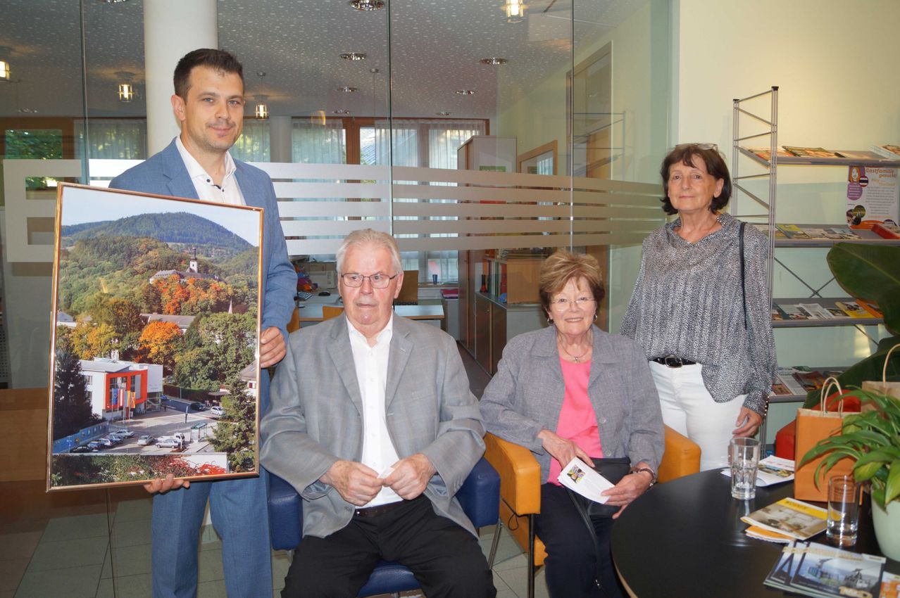 Vier Personen in einem Büro mit einem Landschaftsbild. Zwei Männer und zwei Frauen. Ein Mann hält ein gerahmtes Bild. Die Frauen sitzen auf Stühlen.