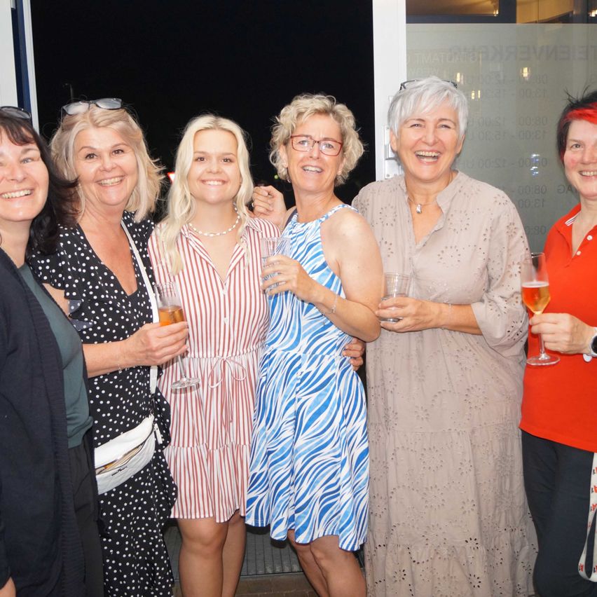 Eine Gruppe von Frauen lächelt und hält Gläser mit Bier und Wein in den Händen. Sie scheinen auf einer Party zu sein.
