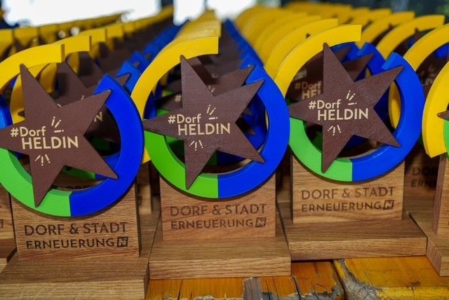 Multiple wooden trophies with blue, yellow, and green bands, featuring a star design, are arranged on a table. The trophies have brown bases with the text Dorf and Stadt Erneuerung.