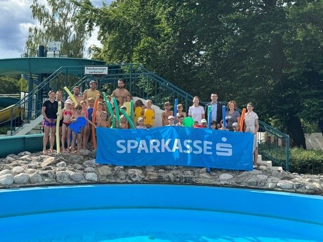 A group of people, adults and children, stand by a pool with a blue banner reading 'SPARKASSE'. They are holding pool toys and wearing swimwear. Behind them, there's a slide and a staircase.