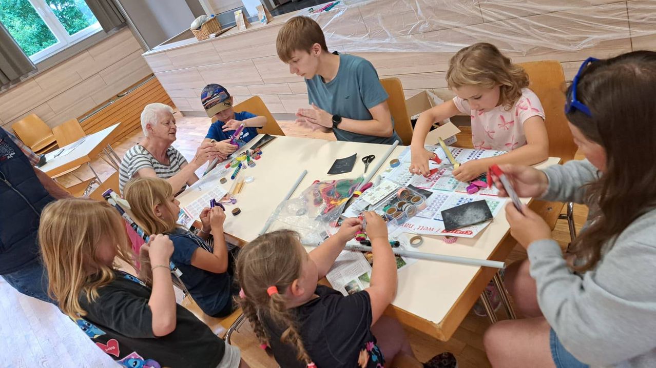 A group of children and an elderly woman are engaged in a crafting activity at a table, surrounded by various art supplies.