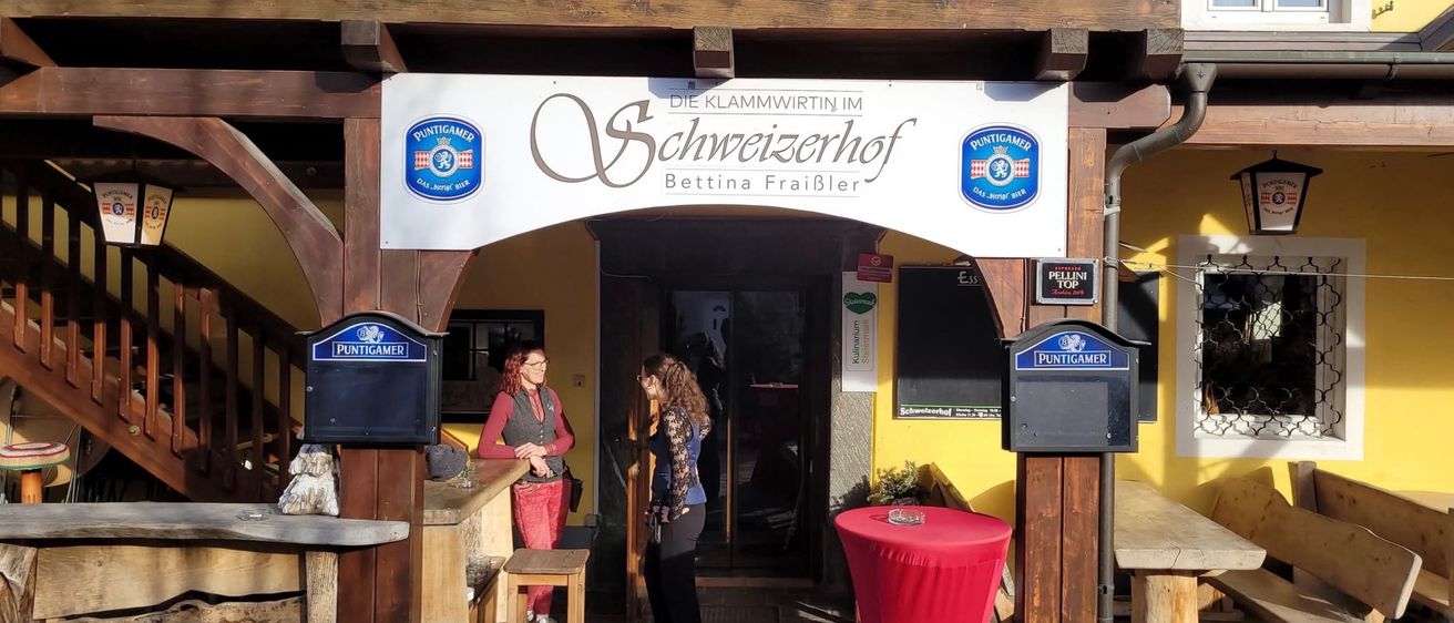 Zwei Frauen stehen vor einer Bar. Ein Schild über ihnen lautet 'Die Klammwirtin im Schweizerhof Bettina FraiBler'. Das Lokal hat einen roten Tisch, eine Holztheke und eine Topfpflanze an der Seite.