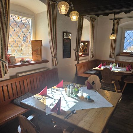 Ein hölzerner Esstisch ist mit Tellern, Besteck und Blumen gedeckt. Der Raum hat Holzbänke, Fenster mit Vorhängen und Lampen, die von der Decke hängen.