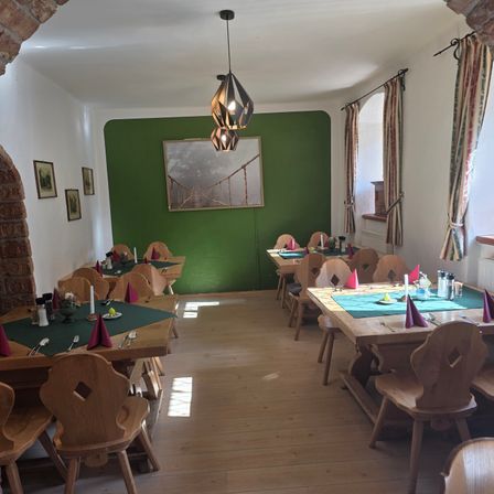 Ein Esszimmer mit Holztischen und -stühlen, einer grünen Wand mit einem Bild und einer Hängelampe. Die Tische sind mit grünen Tüchern, Kerzen und Besteck gedeckt. Der Boden ist aus Holz.