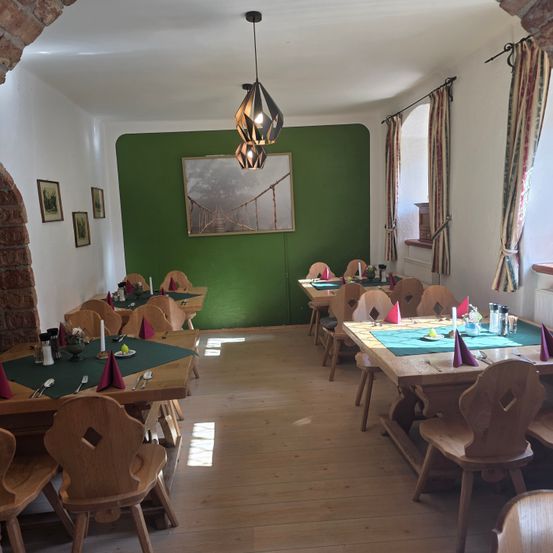 Ein Esszimmer mit Holztischen und -stühlen, einer grünen Wand mit einem Bild und einer Hängelampe. Die Tische sind mit grünen Tüchern, Kerzen und Besteck gedeckt. Der Boden ist aus Holz.