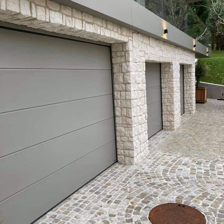 Eine moderne Garage mit drei Türen, einer Steinmauer und einem gepflasterten Weg, mit einer Pflanze in einem Holztopf und Lichtern auf dem Dach.
