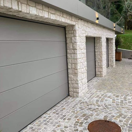 Eine moderne Garage mit drei Türen, einer Steinmauer und einem gepflasterten Weg, mit einer Pflanze in einem Holztopf und Lichtern auf dem Dach.