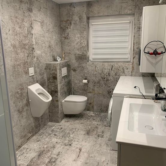 Ein Badezimmer mit modernem Design, ausgestattet mit einem Urinal, einer Toilette, einer Waschmaschine, einem Waschbecken und einem Fenster mit weißen Rolläden.