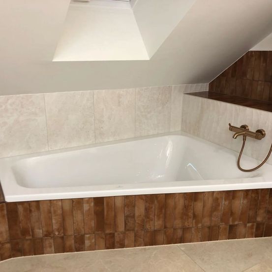 Eine leere Badewanne in einem Dachgeschossbadezimmer mit einer Holzdecke, weißen Wänden und braunen Fliesen. Die Badewanne hat einen weißen Rand und einen goldfarbenen Wasserhahn.