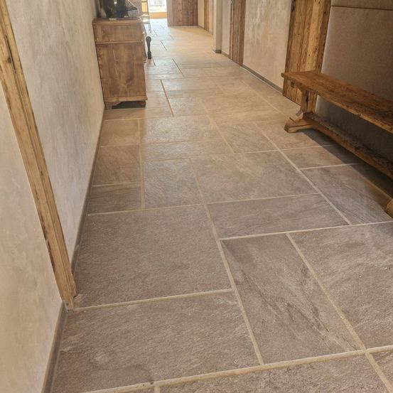 Ein schmaler Flur mit Steinfliesen führt zu einem Raum mit Holztüren. Eine Holzbank befindet sich auf der rechten Seite. Ein Holzschrank mit Gegenständen steht links.