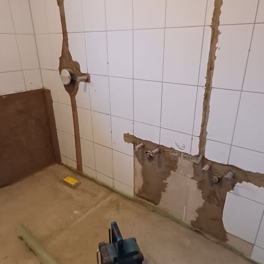 Ein unfertiges Badezimmer mit weißen Fliesen, teilweise entfernten Fliesen und freiliegenden Rohren. Ein gelbes Objekt liegt auf dem Boden und ein Werkzeug ist auf der rechten Seite platziert.