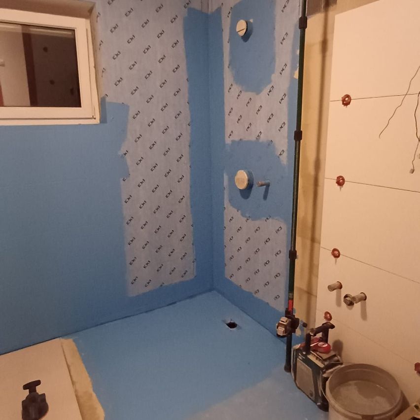 Ein Badezimmer im Bau mit blauen Wänden und Boden. Es gibt ein kleines Fenster, ein Loch und Rohre. Ein Eimer steht auf dem Boden.