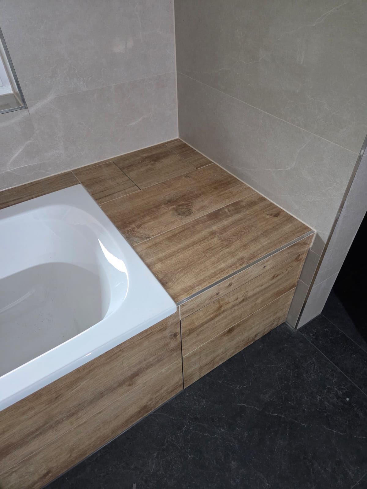 A modern bathroom with a white bathtub and wooden floor next to a wall with light-colored tiles. The bathtub has a smooth surface.