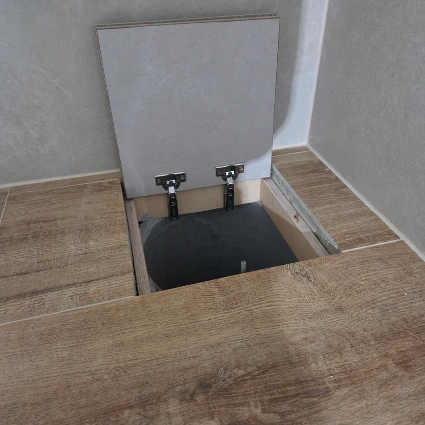 An open access hatch is in the middle of a wooden floor. It has a metal frame with two hinges. The hatch is closed, and the floor is made of wood.