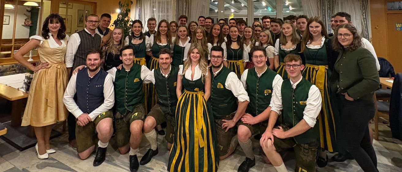 Eine große Gruppe von Menschen in traditioneller Kleidung posiert für ein Foto in einem Raum mit weißen Wänden und einem gefliesten Boden. Einige tragen grüne Westen mit Nummern, andere Kleider. Der Raum ist gut beleuchtet mit Deckenleuchten.