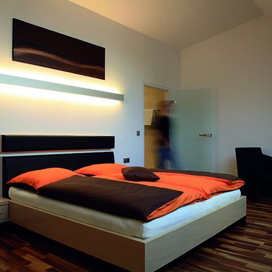 Ein modernes Schlafzimmer mit einem Holzbett, orangefarbenen Kissen und einem schwarzen Kopfteil. Der Raum verfügt über eine Wandleuchte und einen Holzboden. Eine Person ist im Hintergrund verschwommen in der Nähe der Glastür.