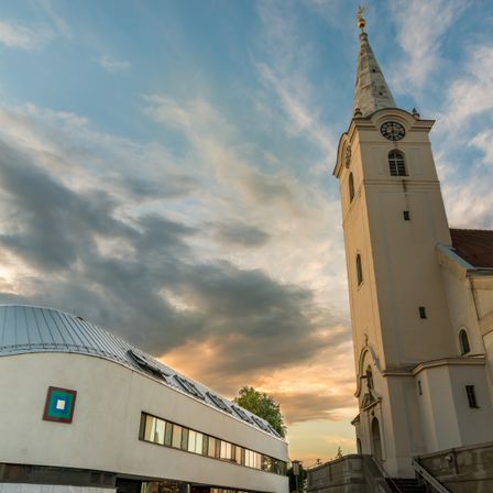 Ein hoher weißer Kirchturm mit einem Dachreiter und einer Uhr steht vor einem bewölkten Himmel. Links davon befindet sich ein modernes Gebäude mit einem quadratischen Logo auf seiner Fassade.