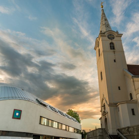 Ein hoher weißer Kirchturm mit einem Dachreiter und einer Uhr steht vor einem bewölkten Himmel. Links davon befindet sich ein modernes Gebäude mit einem quadratischen Logo auf seiner Fassade.