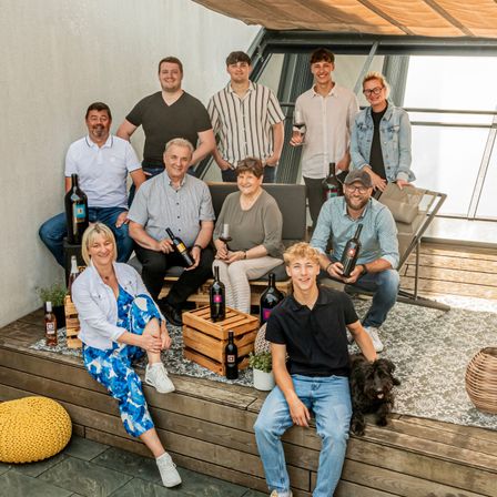 Eine Gruppe von Menschen, möglicherweise eine Familie, posiert für ein Foto auf einer Terrasse. Sie halten Weinflaschen, einige mit Gläsern, und ein Hund sitzt in der Nähe. Holzkisten mit Weinflaschen stehen auf dem Boden.