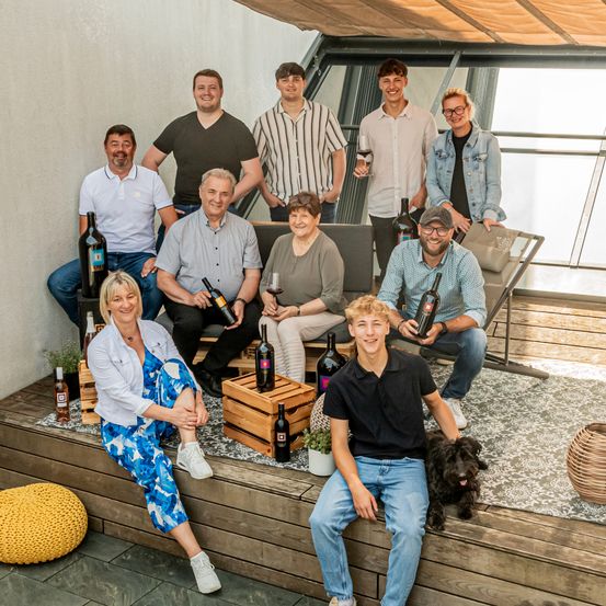 Eine Gruppe von Menschen, möglicherweise eine Familie, posiert für ein Foto auf einer Terrasse. Sie halten Weinflaschen, einige mit Gläsern, und ein Hund sitzt in der Nähe. Holzkisten mit Weinflaschen stehen auf dem Boden.