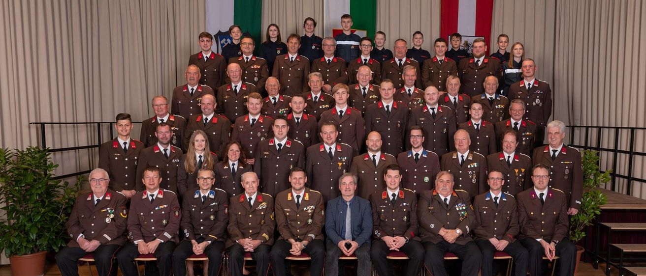 Eine Gruppe von Militärangehörigen in Uniform, sitzend und stehend, mit Flaggen im Hintergrund.