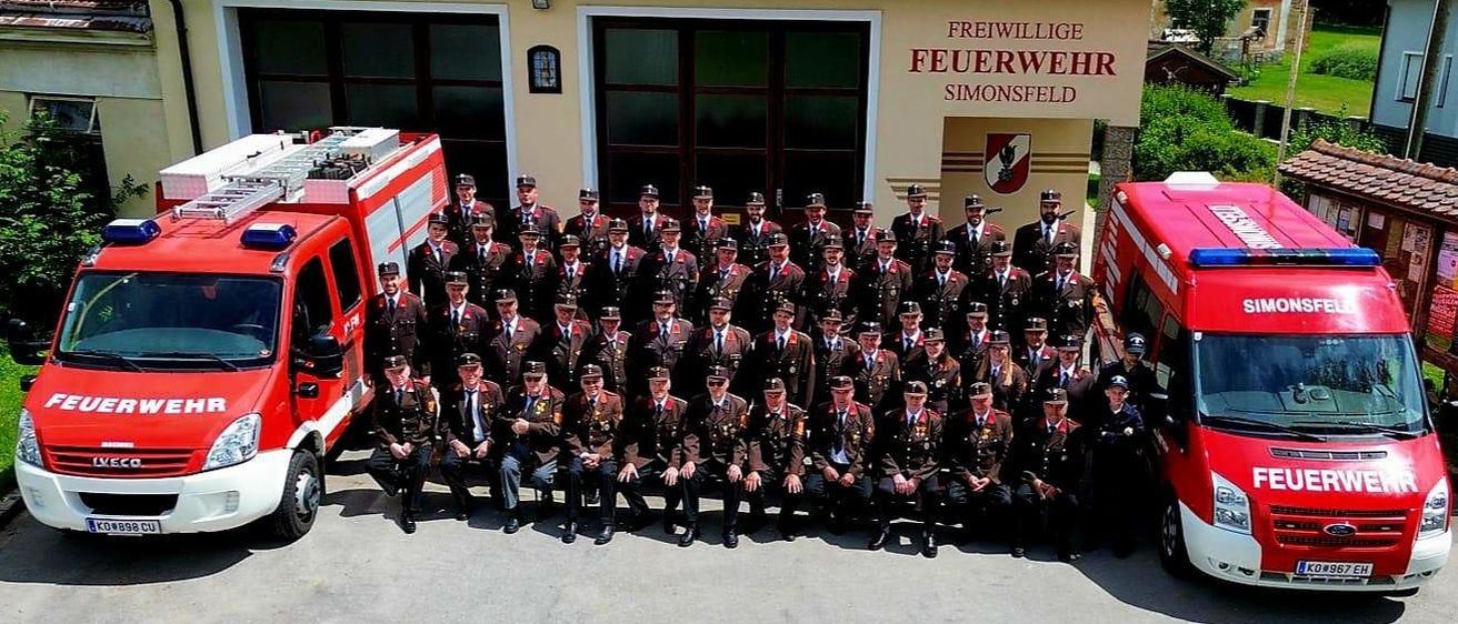 Ein Gruppenfoto von Feuerwehrleuten in Uniform, die vor einem Feuerwehrhaus mit zwei Feuerwehrwagen posieren, wobei der Name des Hauses 'Freiwillige Feuerwehr Simonsfeld' sichtbar ist.